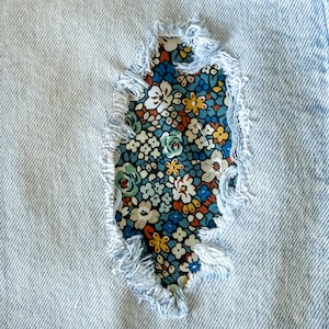 May include: Close-up of light blue denim fabric with a tear, revealing a floral patterned patch. The patch features a colorful array of flowers in shades of blue, white, orange, and yellow. The edges of the tear are frayed.