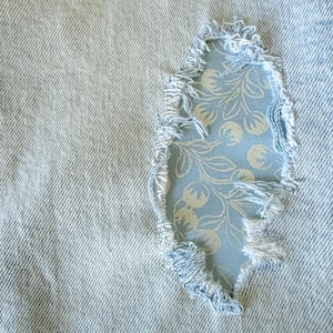 May include: Close-up of light blue denim fabric with a tear, revealing a patch of blue fabric with a white floral pattern. The edges of the tear are frayed, and the patch is visible through the opening. The overall theme is clothing repair or upcycling.