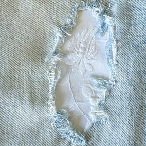 May include: Close-up of light blue denim fabric with a distressed area revealing a white floral embroidered patch. The edges of the denim are frayed, adding a vintage aesthetic to the design.