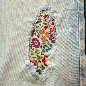 May include: Close-up of light blue denim fabric with a distressed patch. The patch is a floral fabric with red, purple, and yellow flowers. The edges of the denim are frayed, and the patch is sewn in place.