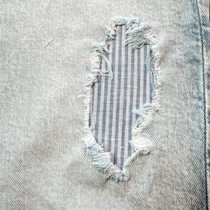 Puede incluir: Primer plano de tela vaquera azul claro con un parche desgastado. El parche revela una tela a rayas azules y blancas debajo, con bordes deshilachados. El denim tiene una apariencia texturizada.