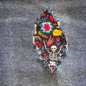 Puede incluir: Un parche de mezclilla con un borde deshilachado, con una inserción de tela negra. La inserción muestra un diseño floral y esquelético colorido. El esqueleto tiene los brazos levantados y las flores son rojas, azules y amarillas.
