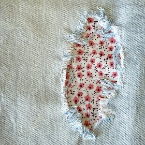 May include: Close-up of light blue denim fabric with a floral patch. The patch is a rectangle of white fabric with a pattern of small pink flowers and green stems. The edges of the patch are frayed with light blue threads.