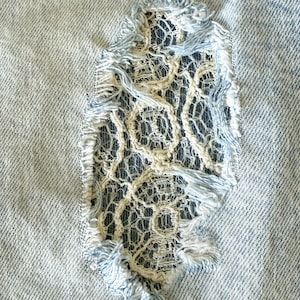 May include: Close-up of distressed denim fabric with a torn section revealing a cream-colored lace insert. The denim is light blue with frayed edges around the tear, creating a textured contrast.
