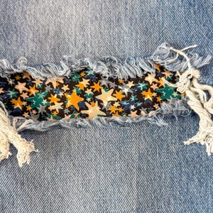 May include: Close-up of distressed denim jeans with a star-patterned fabric patch. The patch features a black background with yellow, teal, and white stars. Frayed edges and light blue denim create a textured, casual look.