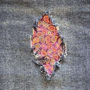 May include: Close-up of a denim fabric with a tear, patched with a paisley pattern fabric. The patch is a diamond shape, showcasing a vibrant mix of colors including red, orange, and green on a purple background.