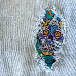 Peut inclure: Gros plan sur un tissu denim bleu clair avec un patch décoratif. Le patch présente un motif de tête de mort mexicaine colorée avec des accents bleus, jaunes et orange. Les bords du patch sont effilochés.