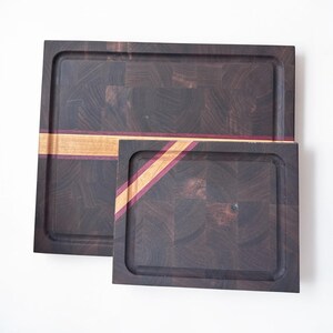 May include: Two rectangular end-grain wooden cutting boards featuring a dark brown checkered pattern. The larger board has a horizontal light wood and purple inlay, while the smaller board features a diagonal light wood and purple inlay.