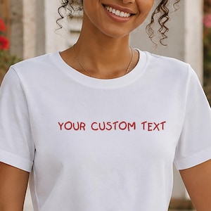 May include: A white short-sleeved t-shirt featuring the text YOUR CUSTOM TEXT printed in red on the chest. The shirt has a crew neckline and a standard fit, constructed from a smooth fabric, providing a clean and simple appearance for daily wear.