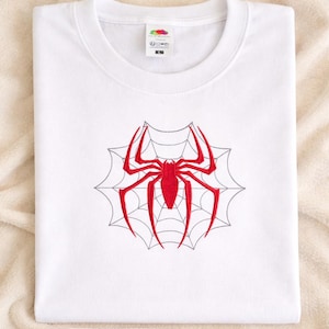 Op de afbeelding: Een wit T-shirt met korte mouwen en een centraal geborduurd ontwerp van een felrode spin over een dun, zwart gestikt spinnenwebpatroon. Het kledingstuk heeft een ronde hals en een zichtbaar label met maat L bij de binnenkraag.