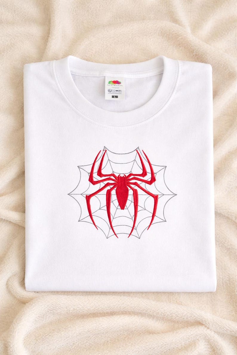 Op de afbeelding: Een wit T-shirt met korte mouwen en een centraal geborduurd ontwerp van een felrode spin over een dun, zwart gestikt spinnenwebpatroon. Het kledingstuk heeft een ronde hals en een zichtbaar label met maat L bij de binnenkraag.