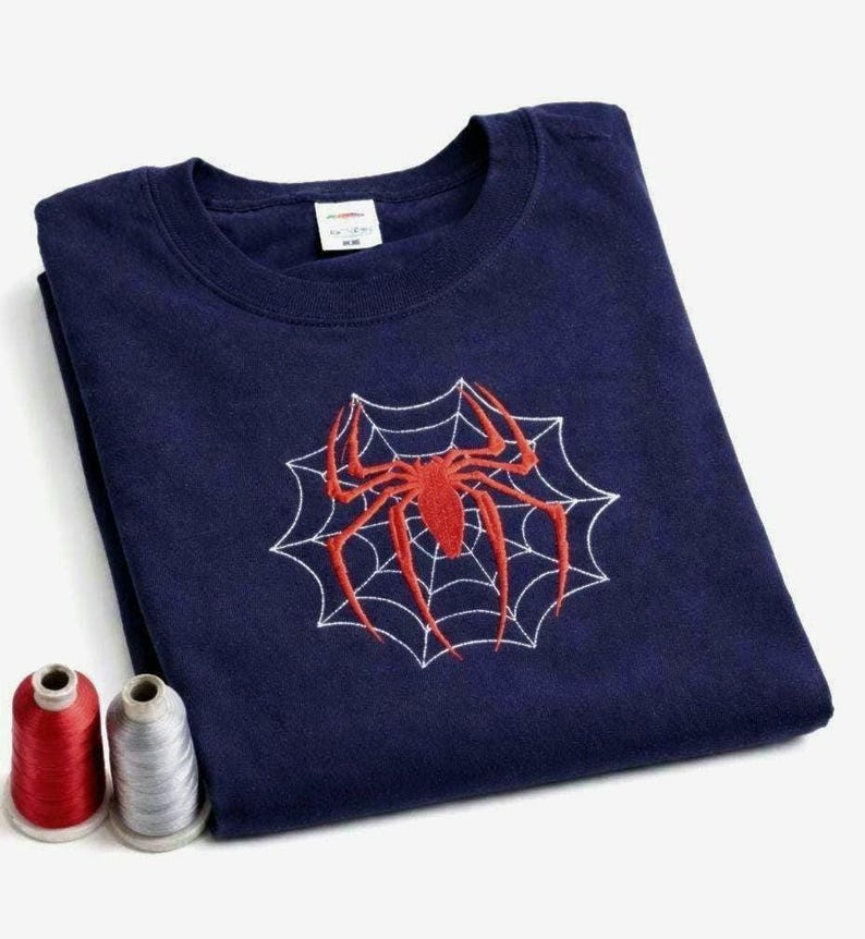 Op de afbeelding: Een marineblauw t-shirt met ronde hals voorzien van een geborduurde rode spin in het midden van een wit gestikt spinnenwebontwerp. Twee kleine klossen garen, &eacute;&eacute;n rood en &eacute;&eacute;n zilverkleurig, liggen linksonder bij het gevouwen kledingstuk.