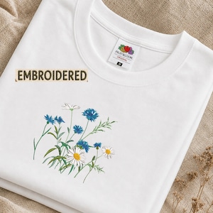 May include: A white t-shirt featuring an embroidered floral design of blue cornflowers and white daisies. The shirt has a crew neckline and a Fruit of the Loom label inside the collar with the size M. The word EMBROIDERED is printed in black.