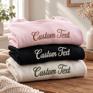 Pode incluir: Uma pilha de três camisolas de gola redonda dobradas em rosa claro, preto e creme. Cada camisola apresenta as palavras Custom Text bordadas em letra cursiva no peito. A camisola rosa tem bordados castanhos, a preta brancos e a creme bordados verde-escuros.