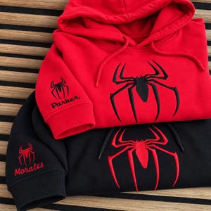 May include: A red hoodie with a black spider emblem and the text Parker on the sleeve, layered over a black hoodie with a red spider emblem and the text Morales on the sleeve. Both hoodies feature matching drawstrings and ribbed cuffs.