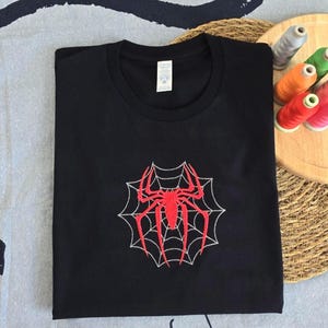 Op de afbeelding: Een zwart t-shirt met korte mouwen en een centraal geborduurd ontwerp van een felrode spin op een wit spinnenweb. Het kledingstuk heeft een ronde hals en een wit label in de nek met daarop zichtbare zwarte tekst.