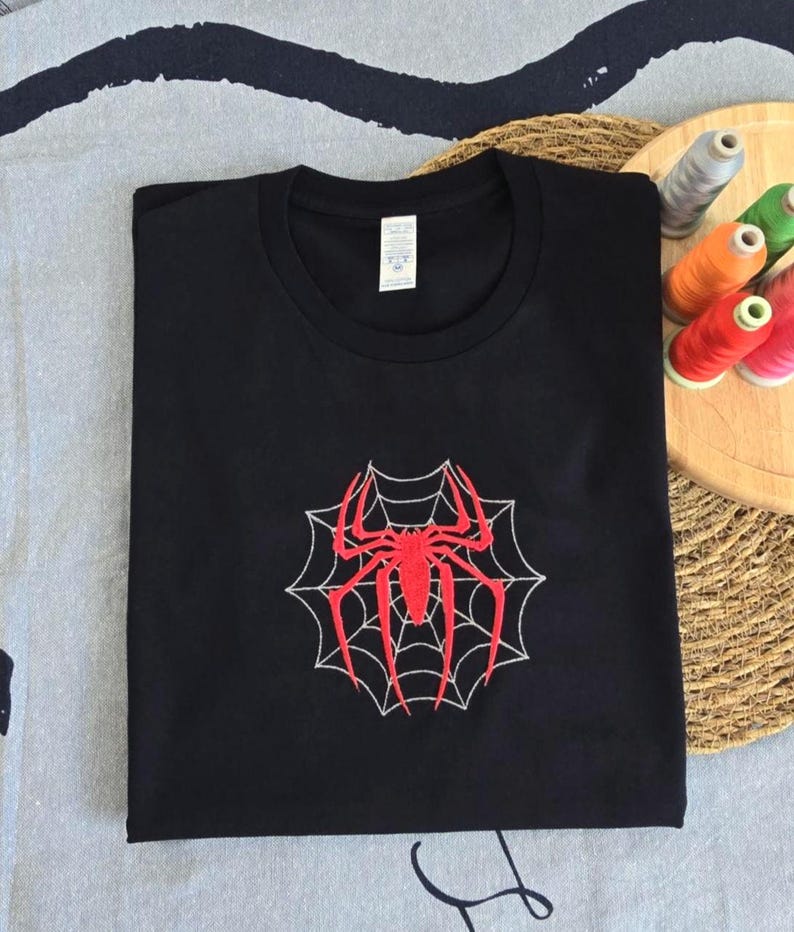 Op de afbeelding: Een zwart t-shirt met korte mouwen en een centraal geborduurd ontwerp van een felrode spin op een wit spinnenweb. Het kledingstuk heeft een ronde hals en een wit label in de nek met daarop zichtbare zwarte tekst.