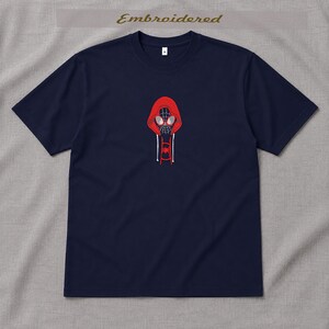 May include: Navy blue t-shirt with an embroidered design of a red hooded figure with a spider-like mask. The design is centered on the chest. The word "Embroidered" is above the design. The shirt has short sleeves.