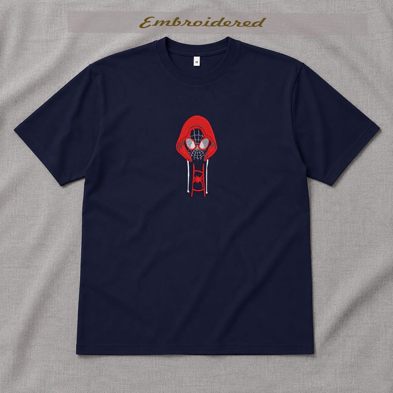 May include: Navy blue t-shirt with an embroidered design of a red hooded figure with a spider-like mask. The design is centered on the chest. The word "Embroidered" is above the design. The shirt has short sleeves.