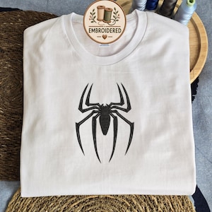 May include: A white short-sleeved t-shirt featuring a black embroidered spider design centered on the chest. The shirt has a round neckline and a circular logo tag at the collar that reads EMBROIDERED with an illustration of two thread spools.