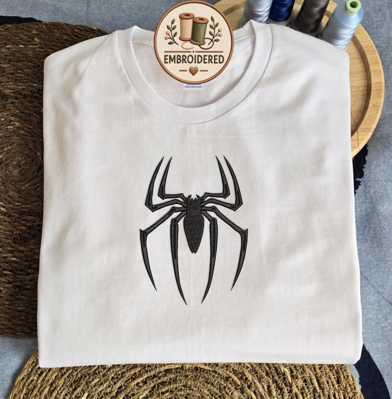 May include: A white short-sleeved t-shirt featuring a black embroidered spider design centered on the chest. The shirt has a round neckline and a circular logo tag at the collar that reads EMBROIDERED with an illustration of two thread spools.