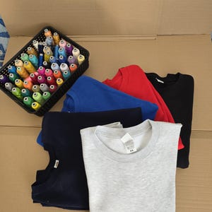 Op de afbeelding: Een collectie van vijf sweaters met ronde hals in lichtgrijs, marineblauw, koningsblauw, rood en zwart, op elkaar gestapeld. Naast de sweaters staat een zwarte rechthoekige plastic mand gevuld met talloze spoelen gekleurd naaigaren.