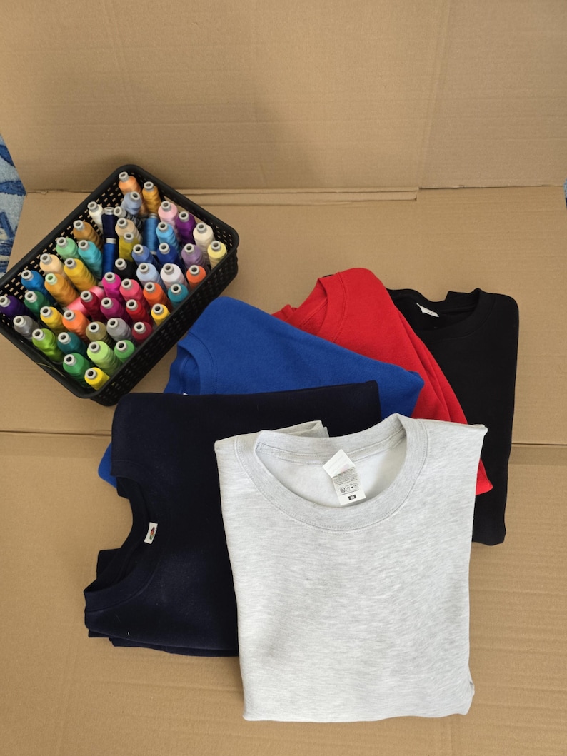 Op de afbeelding: Een collectie van vijf sweaters met ronde hals in lichtgrijs, marineblauw, koningsblauw, rood en zwart, op elkaar gestapeld. Naast de sweaters staat een zwarte rechthoekige plastic mand gevuld met talloze spoelen gekleurd naaigaren.