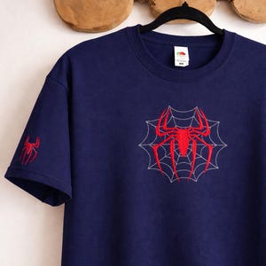 Op de afbeelding: Een marineblauw t-shirt met korte mouwen voorzien van een rode geborduurde spin in het midden van een wit gestikt webontwerp op de borst, met een kleinere rode spin geborduurd op de rechtermouw. Het shirt heeft een ronde hals en een wit label.