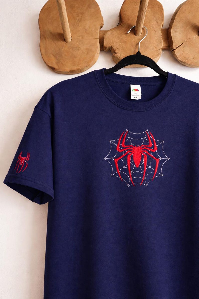 Op de afbeelding: Een marineblauw t-shirt met korte mouwen voorzien van een rode geborduurde spin in het midden van een wit gestikt webontwerp op de borst, met een kleinere rode spin geborduurd op de rechtermouw. Het shirt heeft een ronde hals en een wit label.