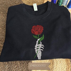 May include: A black crewneck sweatshirt featuring a central embroidered design of a white skeletal hand holding a single red rose with green leaves. The garment has a ribbed neckline and a white tag attached to the inner collar area.
