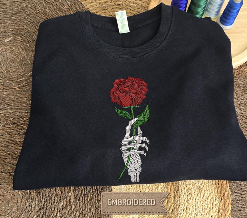 May include: A black crewneck sweatshirt featuring a central embroidered design of a white skeletal hand holding a single red rose with green leaves. The garment has a ribbed neckline and a white tag attached to the inner collar area.