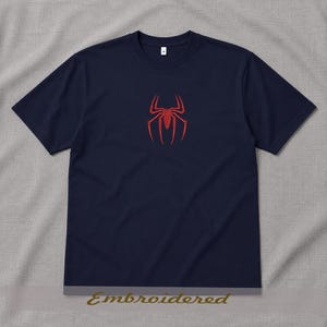 May include: Navy blue t-shirt featuring a red spider emblem on the chest. The shirt has short sleeves and a crew neck. The word "Embroidered" is printed at the bottom of the image. This tee is a casual wear item.