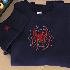 Op de afbeelding: Een marineblauw sweatshirt met ronde hals voorzien van een grote rode geborduurde spin in het midden over een wit gestikt spinnenwebontwerp op de borst, met een kleinere bijpassende rode spin op de mouwboord bij de geribbelde zoom.