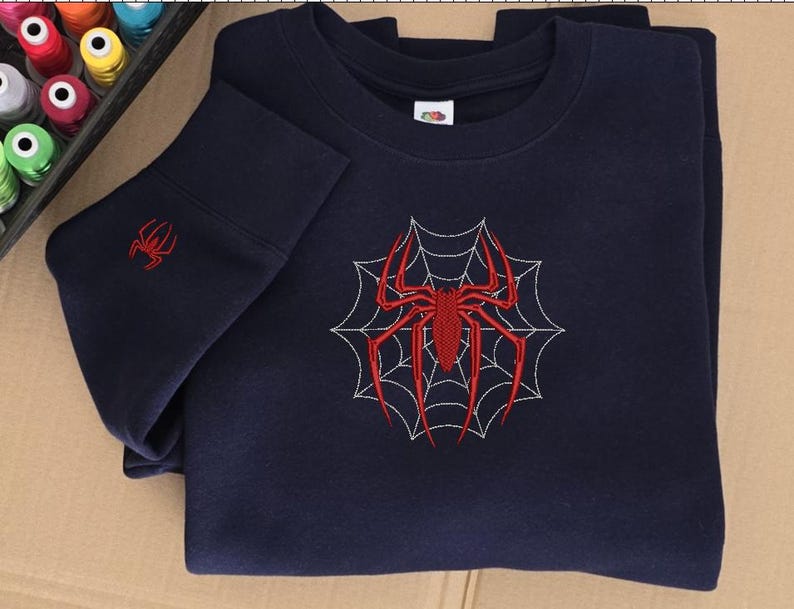 Op de afbeelding: Een marineblauw sweatshirt met ronde hals voorzien van een grote rode geborduurde spin in het midden over een wit gestikt spinnenwebontwerp op de borst, met een kleinere bijpassende rode spin op de mouwboord bij de geribbelde zoom.