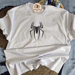May include: A white short-sleeved t-shirt featuring a black embroidered spider emblem centered on the chest. The neckline includes a circular tag with the text EMBROIDERED and a small heart icon. The design is simple and clean.