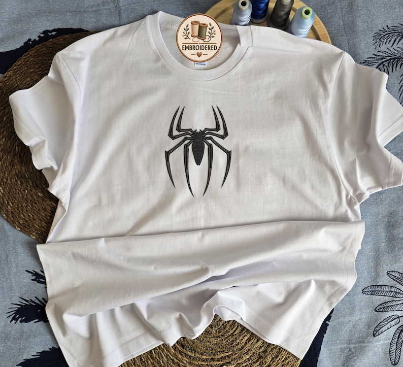 May include: A white short-sleeved t-shirt featuring a black embroidered spider emblem centered on the chest. The neckline includes a circular tag with the text EMBROIDERED and a small heart icon. The design is simple and clean.