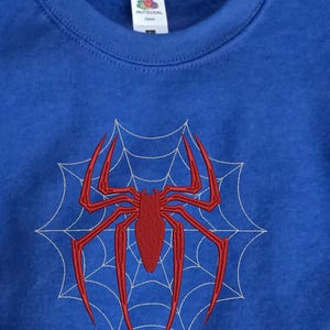 Op de afbeelding: Een koningsblauw sweatshirt met ronde hals voorzien van een groot, centraal rood geborduurd spinnenontwerp over een wit gestikt spinnenwebpatroon. Een kleinere bijpassende rode spinnenborduursel is zichtbaar op de mouwboord.