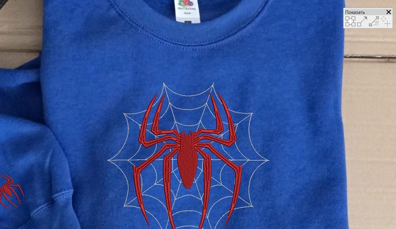 Op de afbeelding: Een koningsblauw sweatshirt met ronde hals voorzien van een groot, centraal rood geborduurd spinnenontwerp over een wit gestikt spinnenwebpatroon. Een kleinere bijpassende rode spinnenborduursel is zichtbaar op de mouwboord.