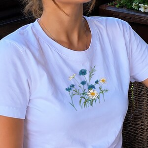 May include: A white short-sleeved t-shirt featuring a central graphic of blue cornflowers and white daisies with green stems. A single blue cornflower graphic is also printed on the right sleeve. The shirt has a crew neckline and a straight hem.