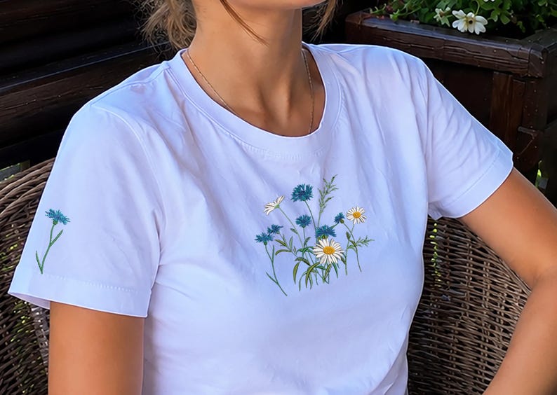 May include: A white short-sleeved t-shirt featuring a central graphic of blue cornflowers and white daisies with green stems. A single blue cornflower graphic is also printed on the right sleeve. The shirt has a crew neckline and a straight hem.