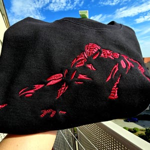 May include: A black crewneck sweatshirt featuring a large, detailed red embroidered graphic of a superhero character in a crouching pose, extending an arm forward. The embroidery uses dense red stitching to create the figure against the black fabric.