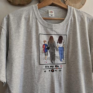 May include: A heather gray t-shirt featuring an embroidered square graphic of three people walking away, with the text "It's my life" and a music player interface icon below the image. The embroidery includes a child in a blue and red jersey.