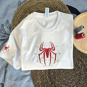 Op de afbeelding: Een wit T-shirt met korte mouwen voorzien van een grote rode geborduurde spin in het midden op een zwart gestikt webontwerp. Een kleinere rode spin is op de linkermouw geborduurd. Het shirt heeft een ronde hals en een wit label.