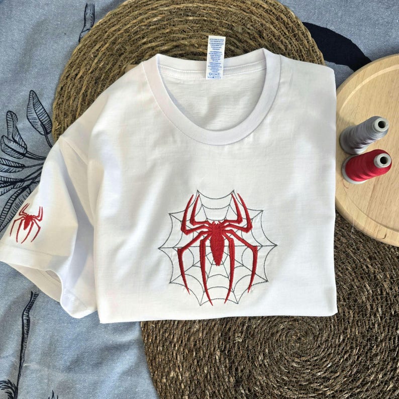 Op de afbeelding: Een wit T-shirt met korte mouwen voorzien van een grote rode geborduurde spin in het midden op een zwart gestikt webontwerp. Een kleinere rode spin is op de linkermouw geborduurd. Het shirt heeft een ronde hals en een wit label.