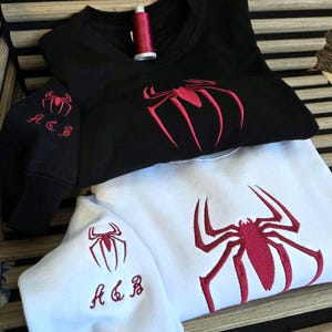 May include: A black sweatshirt and a white sweatshirt, each featuring a large red embroidered spider design on the chest and a smaller spider with the letters A, C, and B embroidered on the sleeve. A spool of red thread sits on the black sweatshirt.
