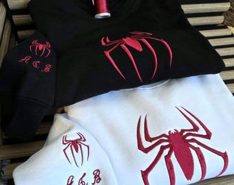 May include: A black sweatshirt and a white sweatshirt, each featuring a large red embroidered spider design on the chest and a smaller spider with the letters A, C, and B embroidered on the sleeve. A spool of red thread sits on the black sweatshirt.