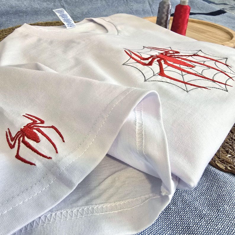Op de afbeelding: Een wit t-shirt met rood geborduurde spinnenmotieven. E&eacute;n rode spin is op de onderkant van de mouw gestikt, terwijl een grotere rode spin centraal in een zwart gestikte spinnenwebafbeelding op de borst van het kledingstuk is geplaatst.