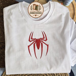May include: A white short-sleeved t-shirt featuring a red embroidered spider design on the chest. A circular logo tag near the neckline displays the text EMBROIDERED with an illustration of two spools of thread and a small heart symbol.