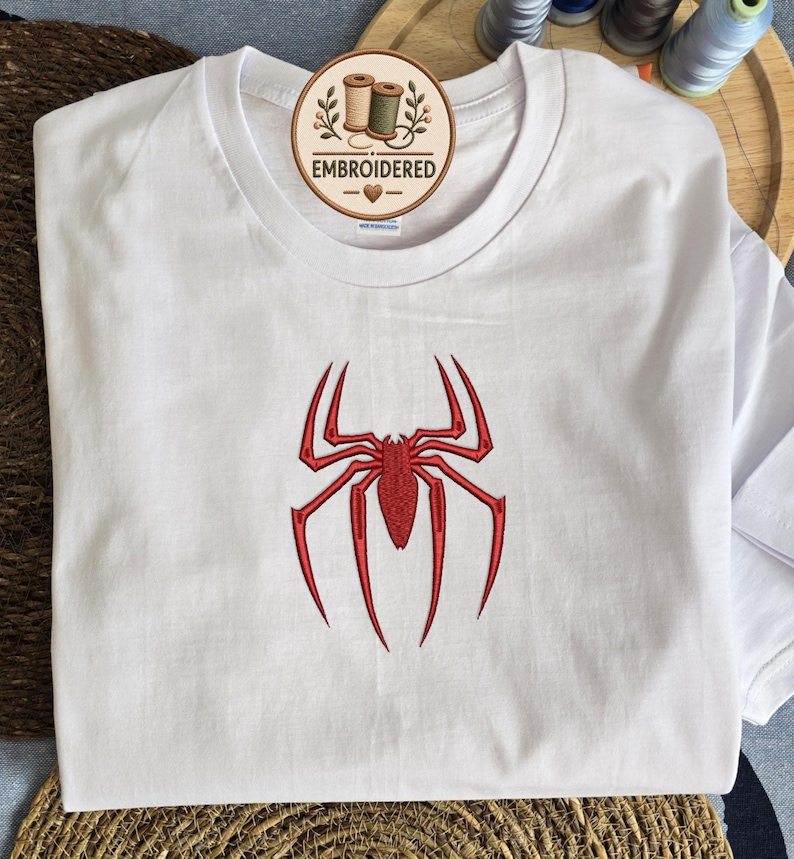 May include: A white short-sleeved t-shirt featuring a red embroidered spider design on the chest. A circular logo tag near the neckline displays the text EMBROIDERED with an illustration of two spools of thread and a small heart symbol.