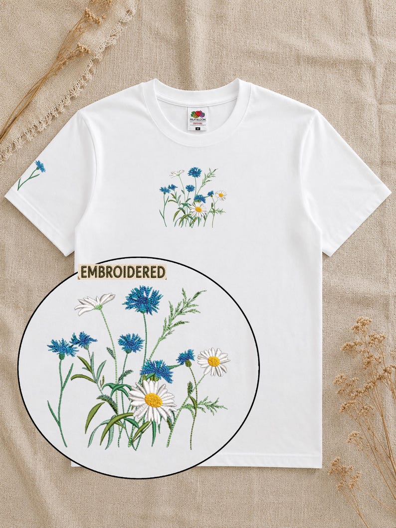 May include: A white short-sleeved t-shirt featuring a central embroidered design of blue cornflowers and white daisies with green stems. A circular inset shows a detailed view of the floral embroidery with the text EMBROIDERED above it.